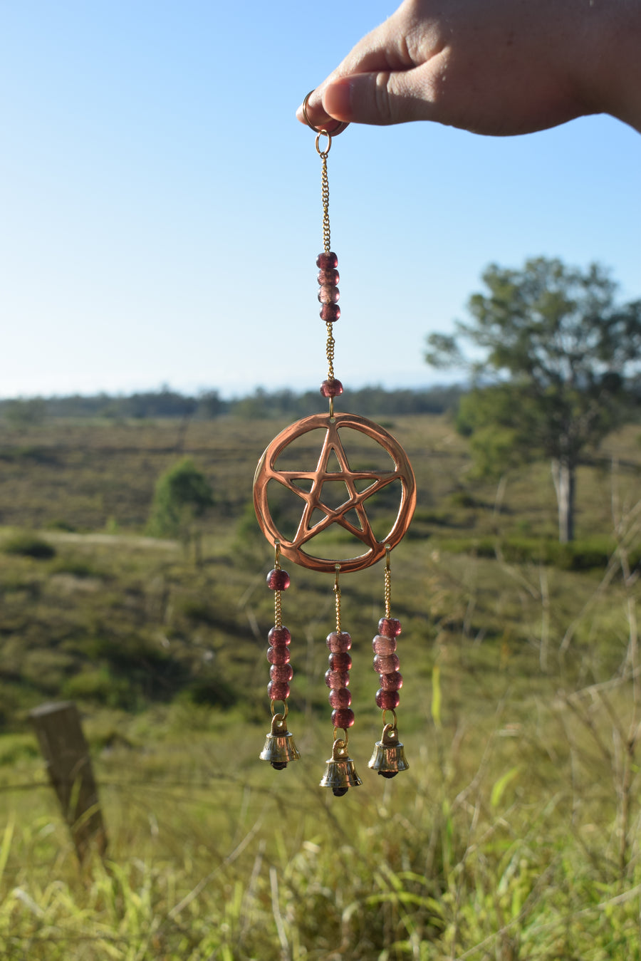 A grape-purple beaded bronze pentagram pentacle metal wind chime is held by a hand with rolling fields and blue sky in the background