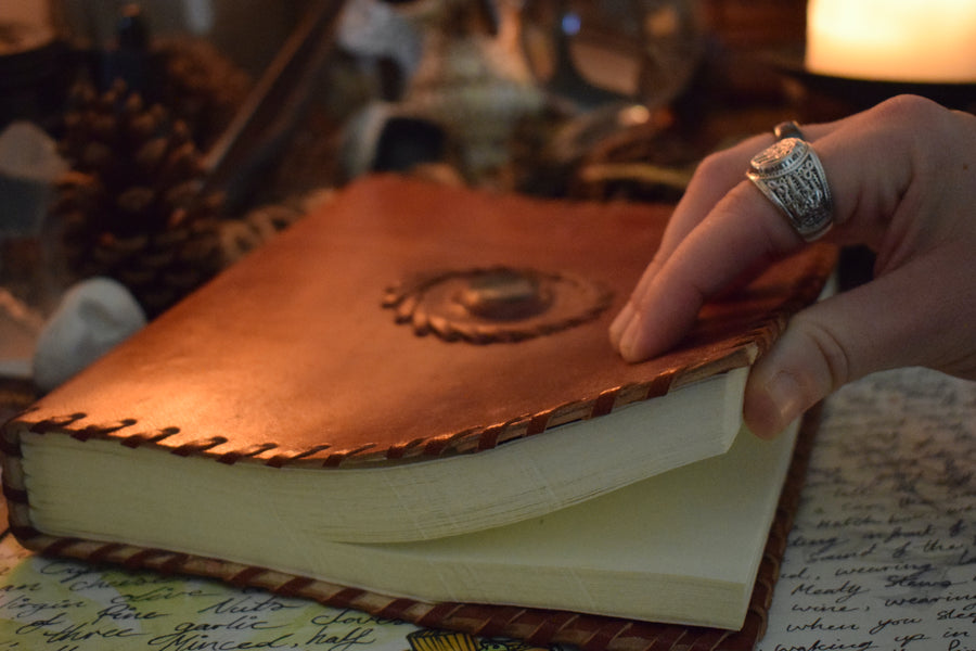 A hand opening a leather-bound book of shadows with a bloodstone crystal sewn into the front cover rests on an altar with candles, tarot cards, crystals and pinecones 