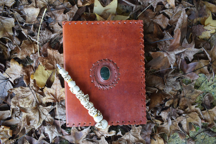 A leather-bound book of shadows with a bloodstone crystal sewn into the front cover rests on a bed of autumn leaves 