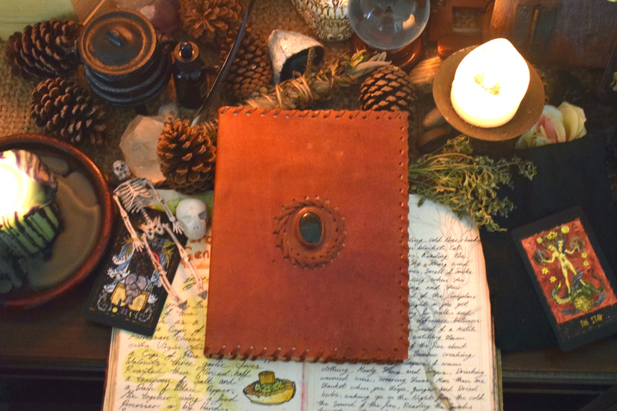 A leather-bound book of shadows with a bloodstone crystal sewn into the front cover rests on an altar with candles, tarot cards, crystals and pinecones 