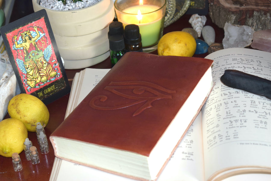 A leather-bound book of shadows with an eye of horus embossed on the cover rests on an open page of hieroglyphs surrounded by crystals, tarot cards, candles and a feather 