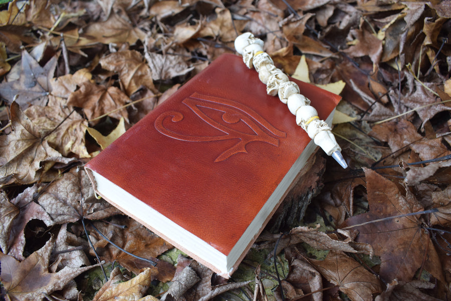 A leather-bound book of shadows with an eye of horus embossed on the cover rests a pen of skulls surrounded by a bed of autumn leaves