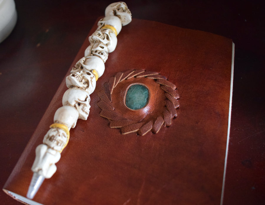 A leather-bound book of shadows with an aventurine crystal inlaid on the cover sits with a pen of skulls