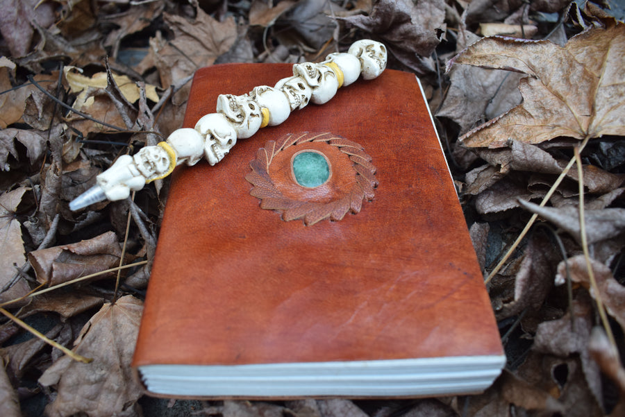 A leather-bound book of shadows with an aventurine crystal inlaid on the cover sits with a pen of skulls on a bed of leaves