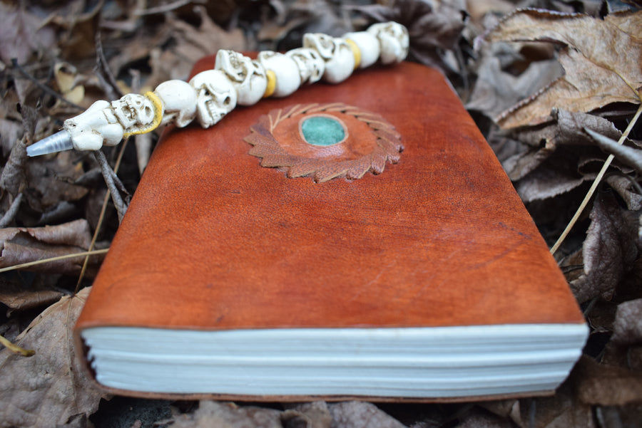 A leather-bound book of shadows with an aventurine crystal inlaid on the cover sits with a pen of skulls on a bed of leaves