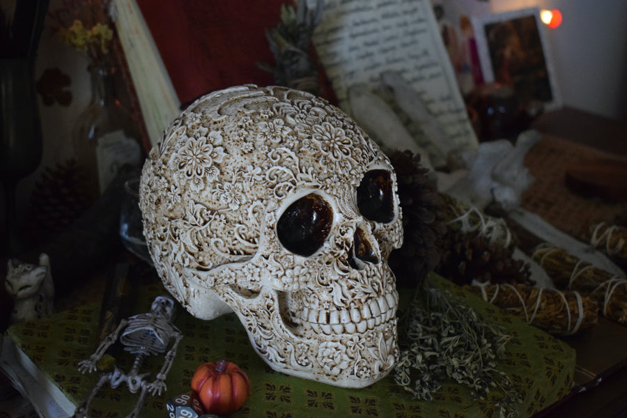 A life sized resin human day of the dead calavera skull with carved floral pattern all over it rests on an altar along with herbs, crystals and a smudge stick.