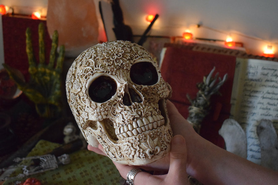 Two hands holding a life sized resin human day of the dead calavera skull with carved floral pattern all over it with an altar in the background.