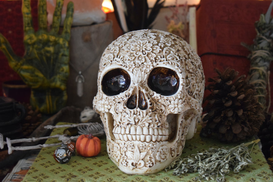 A life sized resin human day of the dead calavera skull with carved floral pattern all over it rests o on an altar along with herbs, pinecone, dice and a skeleton in the background.k.