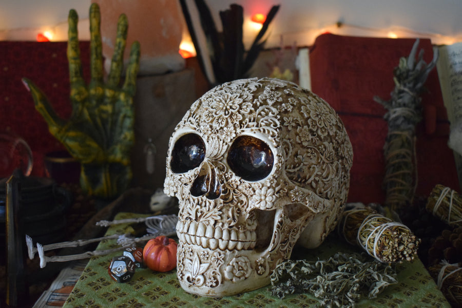 A life sized resin human day of the dead calavera skull with carved floral pattern all over it rests on an open grimoire on an altar along with herbs, crystals and a smudge stick.