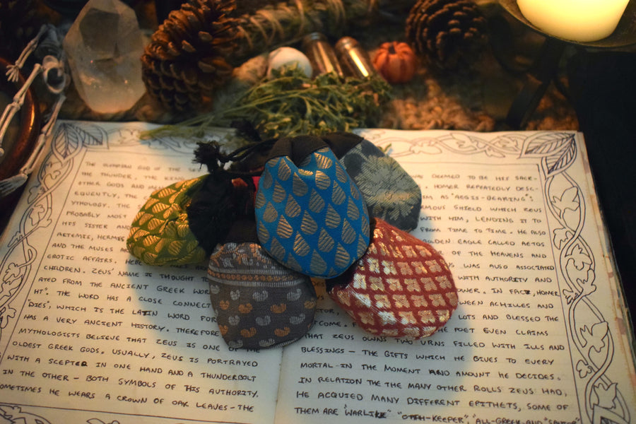 Five different coloured fabric drawstring lucky dip pouches resting on a grimoire with herbs, crystals and candles