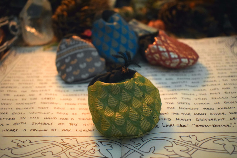 Five different coloured fabric drawstring lucky dip pouches resting on a grimoire with herbs, crystals and candles