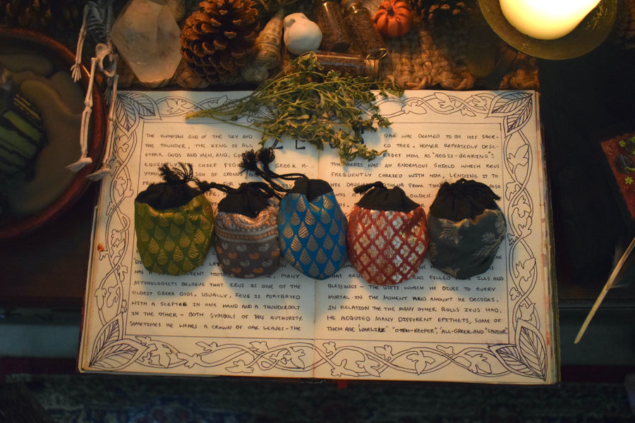 Five different coloured fabric drawstring lucky dip pouches resting on a grimoire with herbs, crystals and candles