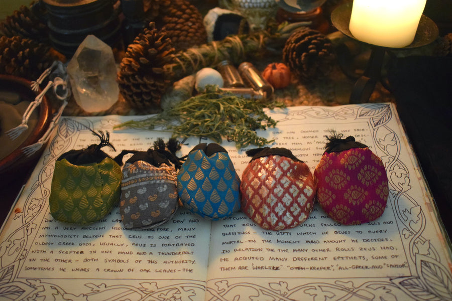 Five different coloured fabric drawstring lucky dip pouches lined up resting on a grimoire with herbs, crystals and candles