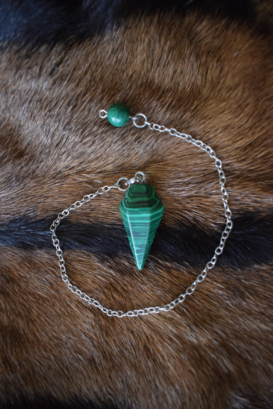 Small malachite point crystal pendulum with chain and bead sitting on goat skin fur