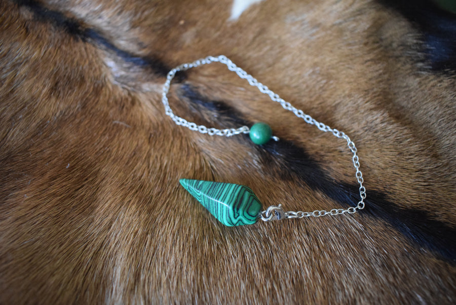 Small malachite point crystal pendulum with chain and bead resting on goat skin fur