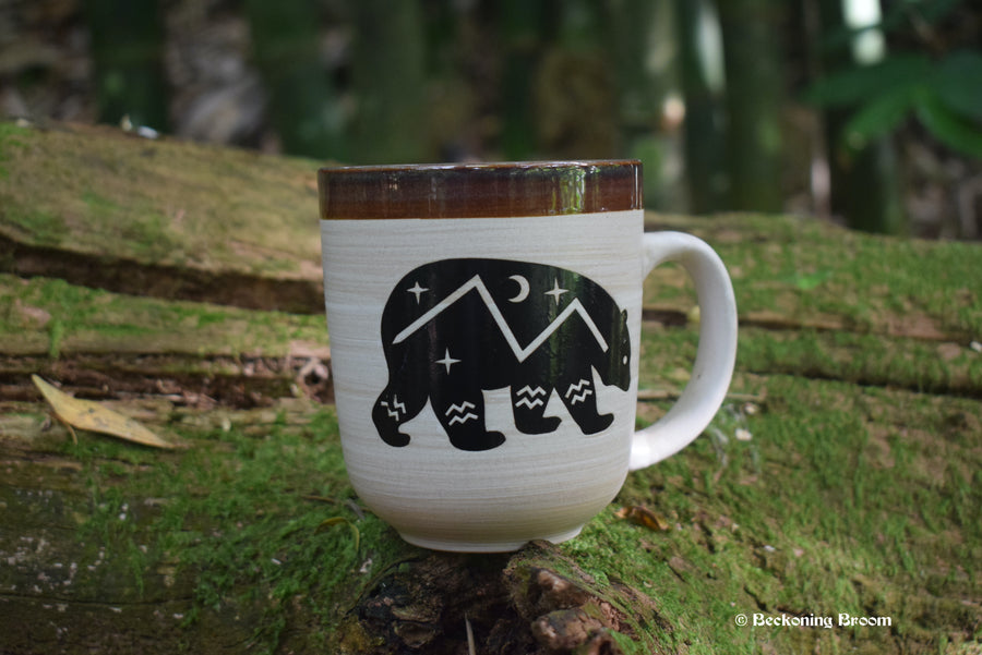 A large mug with a bear depicted on the side with stars, a crescent moon, mountain and river symbols on it rests on a mossy log.