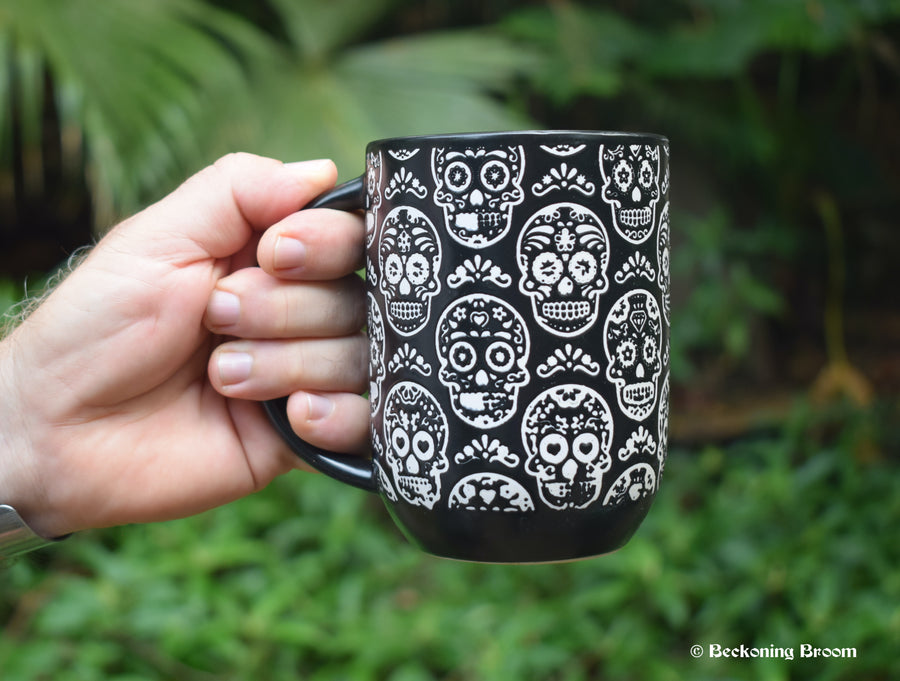 A hand holds a large black mug with white Day of the Dead Calavera skulls all over it. The background is greenery.