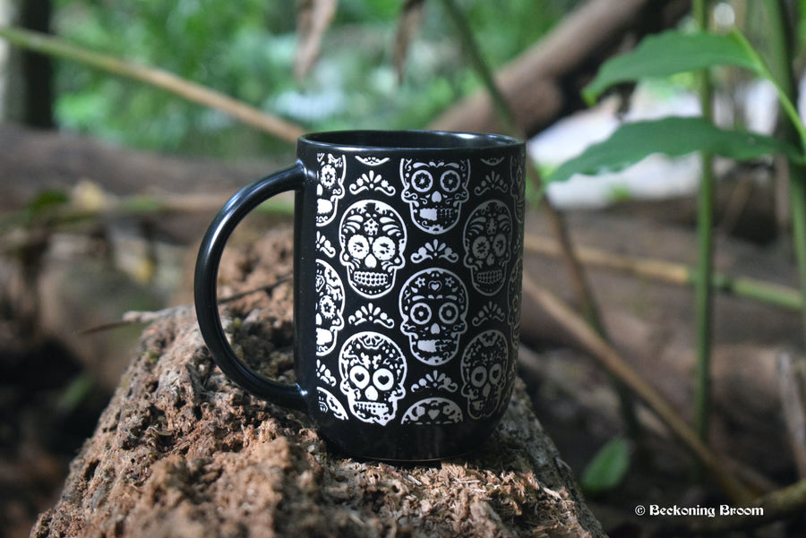 A large black mug with white Day of the Dead Calavera skulls all over it rests on a log.