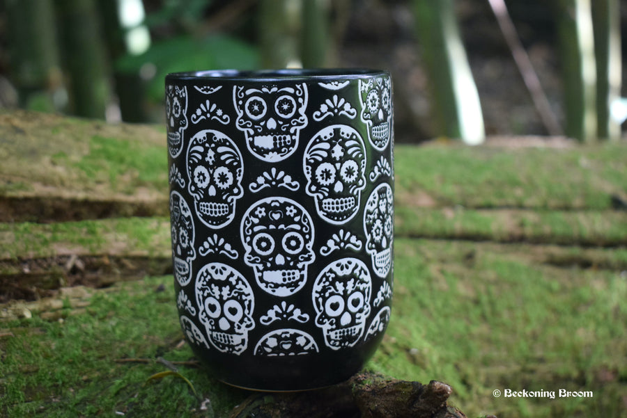 A large black mug with white Day of the Dead Calavera skulls all over it rests on a mossy log.