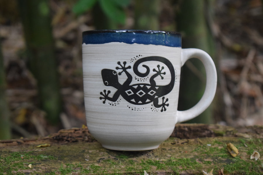A large cream mug with a blue rim and a lizard or gecko on the front rests on a mossy log.