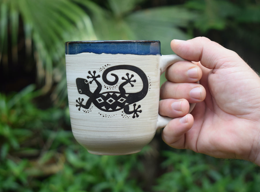A hand holds a large cream mug with a blue rim and a lizard or gecko in black on the front with greenery in the background.