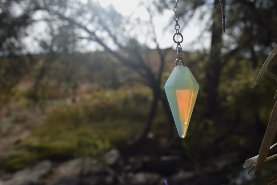 Small opalite point crystal pendulum with chain hanging in the air with trees in background