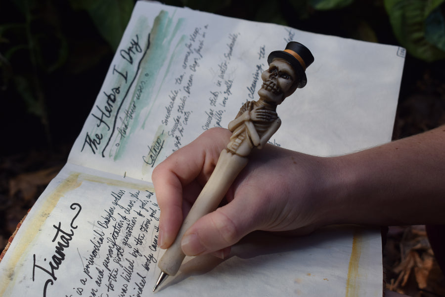 A hand writing in a grimoire with a pen in the shape of a skeleton with arms crossed and a top hat