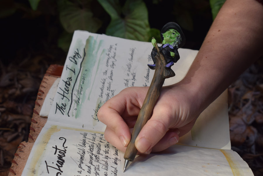 A hand writing in a grimoire with a pen that looks like a piece of wood with a witch riding a broomstick on the end of it