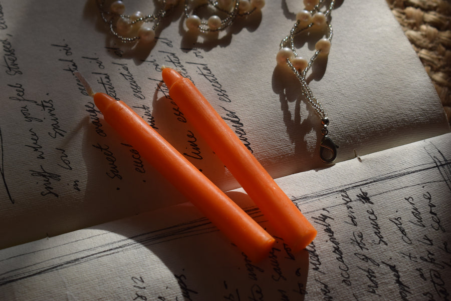 Two orange spell candles rest on an open grimoire with a string of pearls.