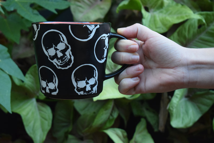 Hand holding black coffee mug with white skulls and orange inside with green plant background