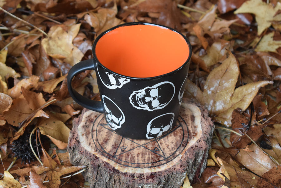 Large black ceramic mug with white skulls and orange inside on pentagram wood block surrounded by dry leaves