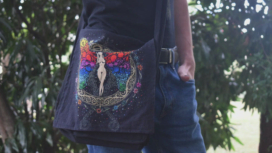 Person in blue jeans and black t-shirt taking book out of rainbow angel, fairy, goddess printed black shoulder bag 