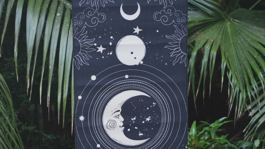 A black fabric tapestry depicting white moons and a sun with a long black fringe hangs with greenery in the background.