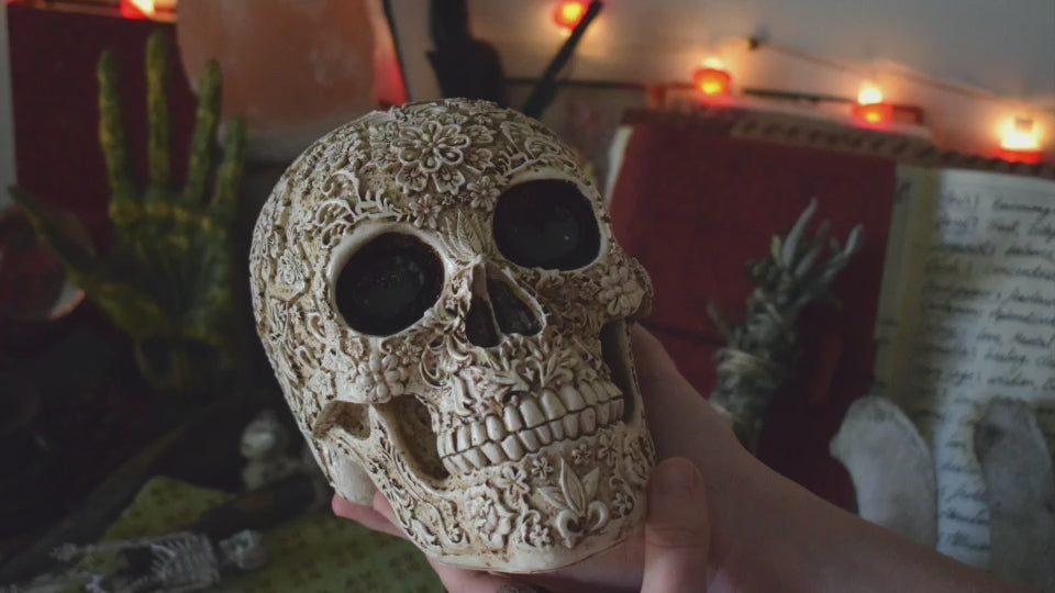 A life sized resin human day of the dead calavera skull with carved floral pattern all over it rests on an altar along with herbs, crystals and a smudge stick.