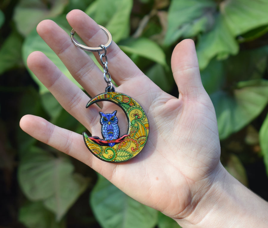 A hand holds a key ring with a resin blue owl perched on a colourful crescent moon on a rock with a leafy background.