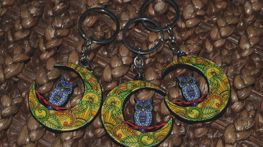 Three key ring with a resin blue owl perched on a colourful crescent moon rests on a paperbark bark background.